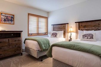A cozy bedroom at Pines Condominiums Two Bedroom features two twin beds with wooden headboards, green comforters, and bear-themed pillows, alongside a rustic dresser and a window with blinds.
