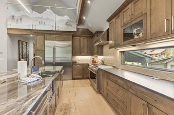 A modern rustic kitchen featuring light wood floors, dark wood cabinetry, a large marble-like island with a sink, stainless steel appliances, and a unique panoramic window with an exterior view.