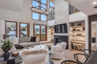 A luxurious and spacious living room at Woodrun Place 56 features high ceilings, a comfortable sectional sofa, and a large fireplace with a mounted TV, all set against a backdrop of snowy mountain views.