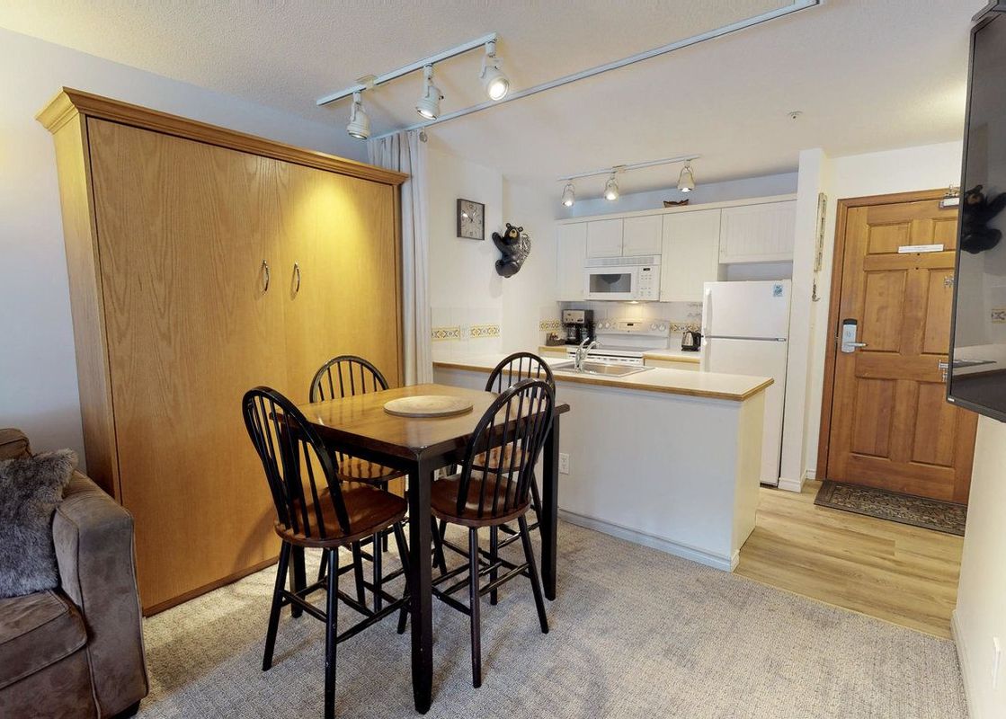 The Aspens on Blackcomb Unit 234 features a dining area with a wooden table and four chairs, a compact kitchen with white appliances and cabinets, and a murphy bed unit, all within an open-concept living space.