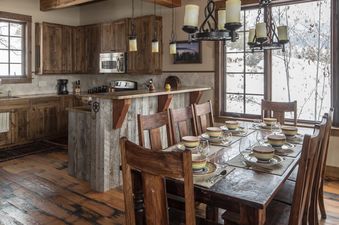 A rustic kitchen and dining area with wooden cabinetry, a breakfast bar, and a dining table set for a meal, with large windows overlooking a snowy landscape.