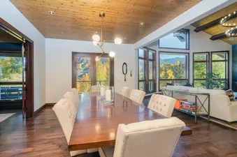 A spacious, upscale dining room in the Evergreen home featuring a long wooden table, tufted white chairs, and windows showing mountain views.