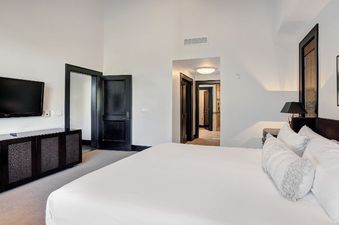 A spacious and modern bedroom with a large bed, wall-mounted TV, dark wood furniture, and artistic decor, with doorways leading to other rooms in the Hummingbird Platinum 3 BR Plus Loft unit.