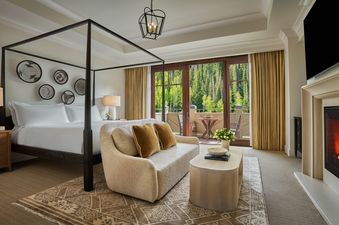 A hotel bedroom featuring a four-poster bed, a sitting area with a sofa, and a balcony overlooking a forest.