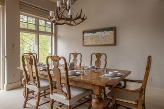 A rustic dining area with a wooden table set for eight, an antler chandelier, and a large window overlooking green trees.