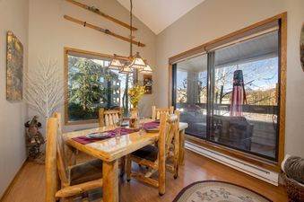 The dining area at Snowblaze C24 features a rustic wooden table set for four, with large windows overlooking trees and a private patio.