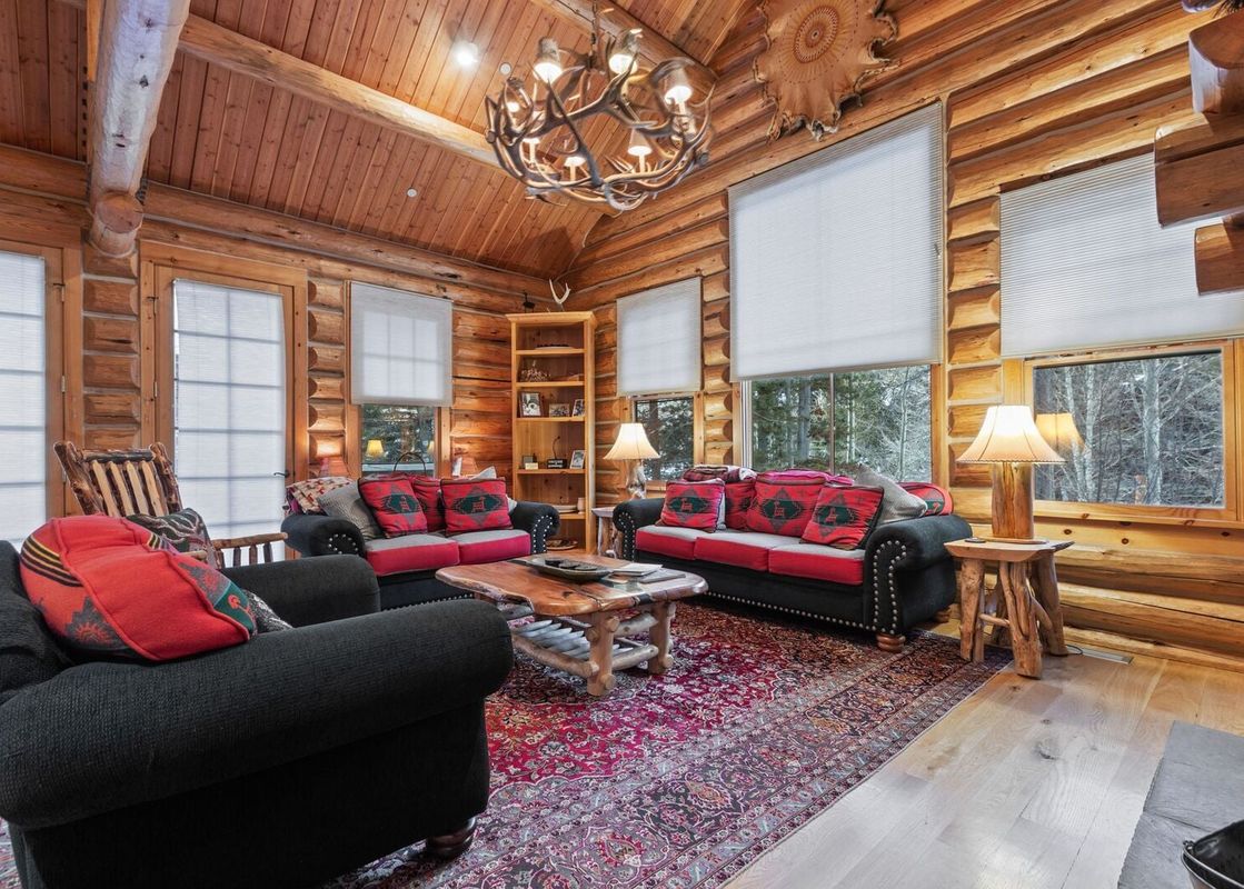 The rustic living room at Obsidian Jackson Hole features log walls, a vaulted wooden ceiling, and a large antler chandelier illuminating two black sofas and an armchair with red pillows, centered around a wooden coffee table and a patterned rug.