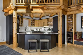 A modern kitchen with a large island and two bar stools, featuring light wood cabinetry, black appliances, and rustic log beam architecture.