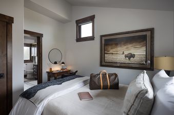 A cozy bedroom in Northern Exposure features a bed with white and gray bedding, a wooden desk with a lamp and mirror, a large framed bison artwork, and an ensuite bathroom visible through an open doorway.