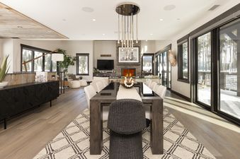 A modern dining room with a long wooden table, white chairs, and a sleek chandelier, extending into a living area with a fireplace and large windows showcasing a snowy outdoor scene.