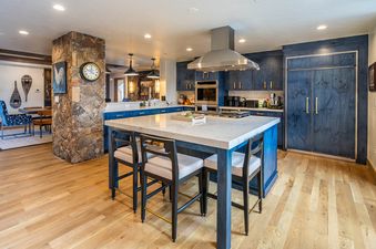 A modern kitchen with a large island, dark blue cabinetry, and stainless steel appliances is shown, featuring a rustic stone pillar with a clock and dining area in the background.