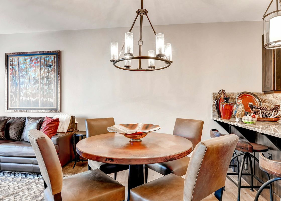 A brown leather sectional sofa with red pillows sits in front of a copper table set with four chairs in a living room with wood floors and a chandelier hanging from the ceiling.