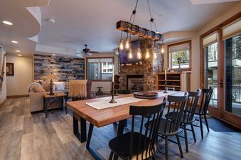 A rustic-chic dining area with a long wooden table, black chairs, and a unique beam chandelier with Edison bulbs, flowing into a cozy living room with a stone fireplace and plush seating at Chateau Chamonix CX326.