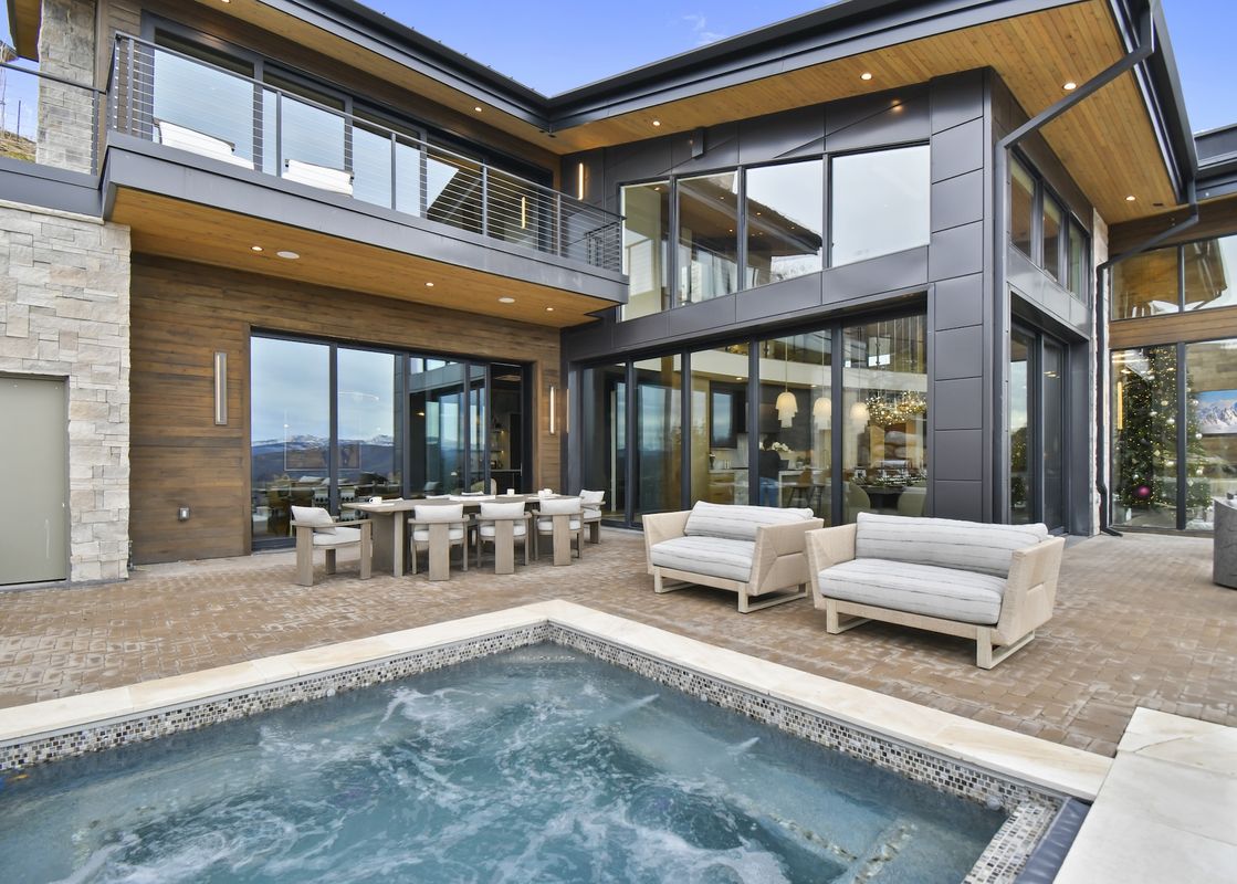 An inviting outdoor patio with a hot tub and seating areas, set against the backdrop of a modern architectural home with large windows and mountain views.