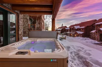 The Tristant 125 hot tub is set against a snow-covered landscape with wooden lodges, illuminated by the vibrant hues of a sunset sky.