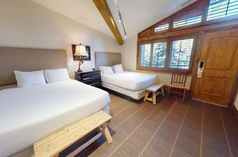 Manor Vail 227 bedroom with two queen beds, a wood bench, a lamp on a side table, and a view out the window of a wooden door and the forest.
