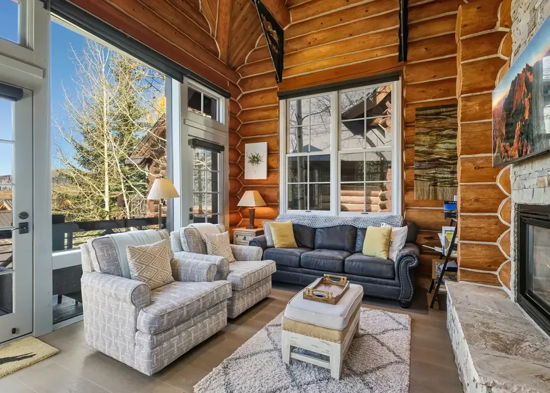 A cozy living room in a log cabin style home, featuring a stone fireplace, plush seating, and large windows with an exterior view of trees and mountains.