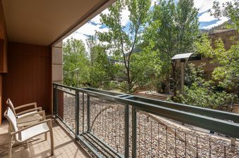 A serene balcony view from North of Nell Unit 2D, featuring patio chairs, a sturdy dark green railing, and a lush outdoor landscape with trees and river stones.