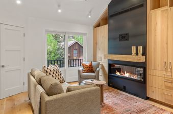 A bright living room features a comfortable sofa and armchair, a modern fireplace, light wood cabinets, and a window with a view of green trees and distant houses.