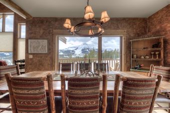 A rustic dining room with a large wooden table, patterned chairs, and a chandelier, offering a spectacular view of snow-capped mountains and pine trees through expansive windows.