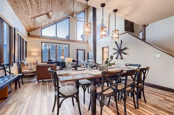 The Ullr Haus dining area features a large wooden table with set places, surrounded by black chairs, under three cylindrical pendant lights, with a view into the living space.