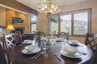A round wooden dining table set for six with white dinnerware and wine glasses, illuminated by a chandelier, with large windows overlooking a snowy mountain landscape.