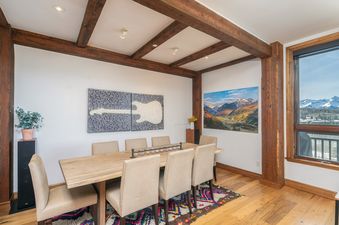 A spacious dining room at See Forever San Sophia 114 3BR 4BA features a long wooden table with eight upholstered chairs, decorative ceiling beams, and two large art pieces, with a window offering mountain views.