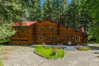 The Apres Chalet is a grand log home surrounded by a dense evergreen forest, featuring a red roof, a gravel driveway, and a small, heart-shaped garden with colorful flowers.