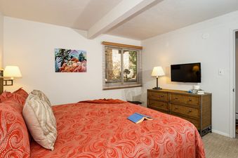 A serene bedroom at Aspen Alps Apartment 101 featuring a bed with a red and white patterned duvet, a vibrant painting, a window with blinds, and a dresser with a TV.