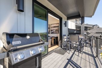 A private outdoor deck featuring a Weber grill, an outdoor dining table with chairs, and a sliding glass door leading to the interior living space, with modern outdoor lighting.