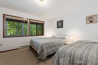 A bright bedroom with two beds, each covered in a gray and white striped comforter, features large windows showcasing a lush green forest outside.