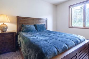 A cozy bedroom at Ranch at Steamboat RA604 featuring a queen-sized bed with blue bedding, a dark wood headboard, a bedside table with a lamp, and a window with an outdoor view.