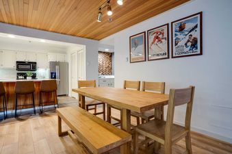 A dining table with a bench and chairs is in the foreground, with a modern kitchen visible in the background, featuring white cabinets and stainless steel appliances.