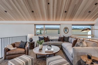 The living room at Havens Haus features a cozy sectional sofa, a leather armchair, a round wooden coffee table, and large windows with a view of the snowy exterior.