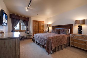 A king-sized bed with an ornate comforter and leather headboard is flanked by stylish lamps in the master bedroom of Chateau Chamonix CX132, featuring a mounted TV, wooden dresser, and a window with curtains.