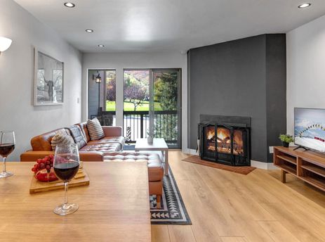 A cozy living room at Snowcrest 216 with a leather sectional, fireplace, television, and a table with red wine and fruit, offering a view of green hills through glass doors.