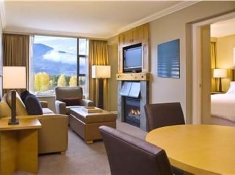 A cozy, sunlit living room at The Westin Resort One Bedroom Suite, with a fireplace, comfortable seating, a dining area, and a large window overlooking a scenic mountain landscape with autumn foliage.
