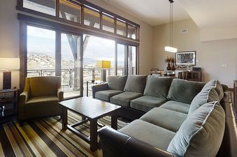 The living area of Trailhead Lodge 4100 features a cozy grey sectional sofa and large windows overlooking a snowy winter landscape, complemented by a unique wooden slat ceiling.
