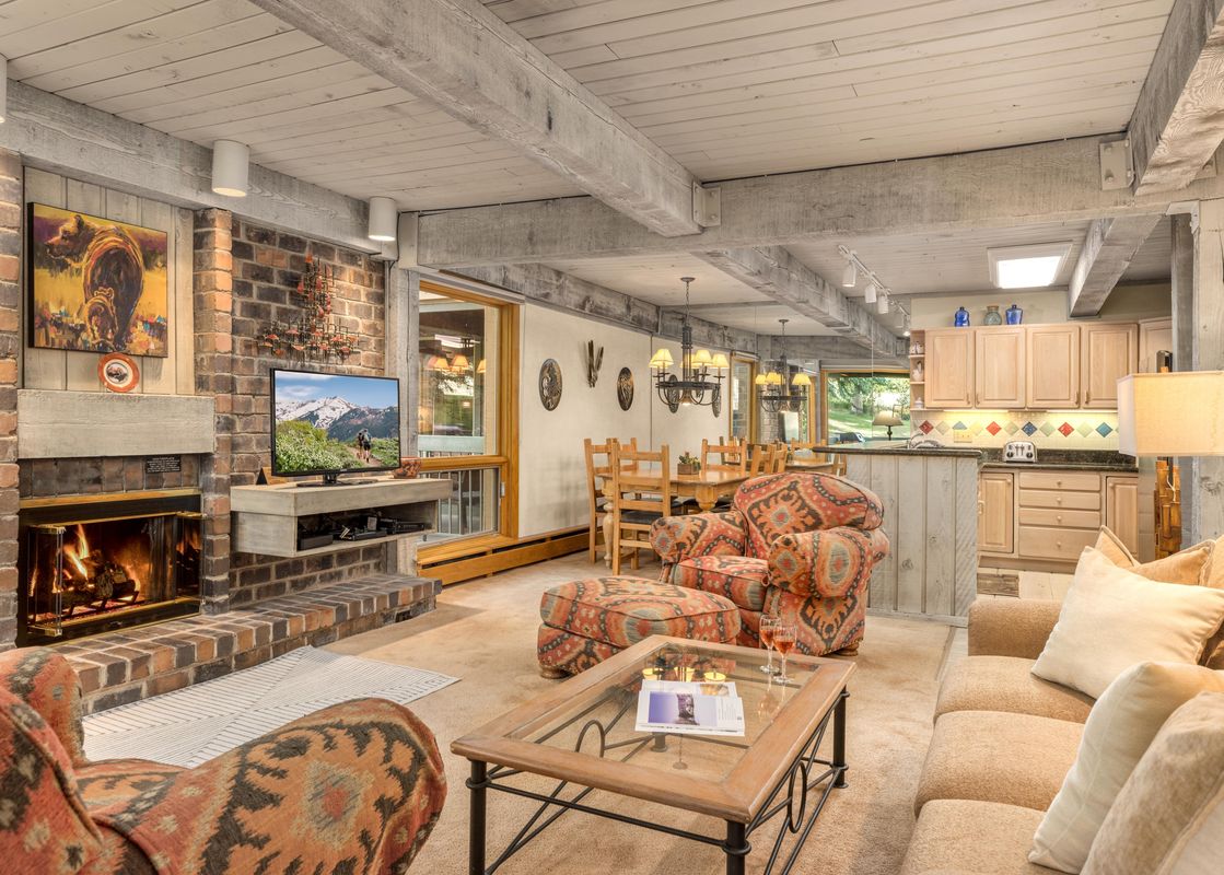 A cozy living room with a brick fireplace, a television displaying a mountain scene, and plush seating, featuring a blend of rustic and comfortable elements.
