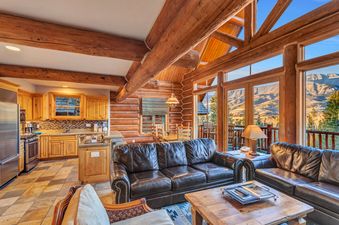 The living room of Mountain Lodge 6 Bedroom Luxury Cabin has a beautiful view of the mountains. It features a large leather couch, a coffee table, and a lamp.