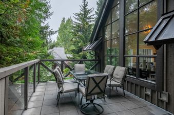 A private balcony at Whistler Pinnacle Ridge 42, featuring a dark-framed glass-top table with six cushioned chairs, all set on a tiled deck with a glass-paneled railing, surrounded by lush green trees and a dark-stained wooden exterior with a large window