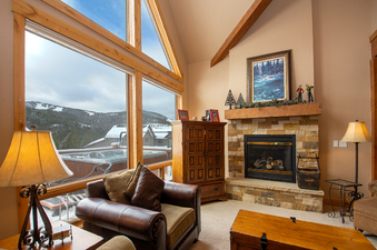 The living area at Antlers at Lakota 406 features a fireplace with a stone facade, comfortable seating, and a large window showcasing a snowy mountain view with a hot tub on the deck.