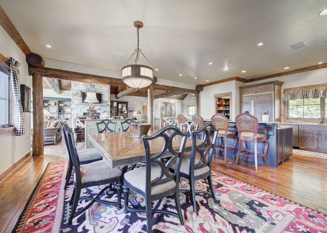 A rustic yet elegant dining room at Peaks View Lodge with a large wooden table, dark chairs, a patterned rug, and a chandelier, leading to an open-concept living room and kitchen.