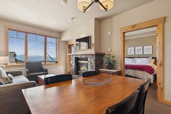 A spacious, open-concept living and dining area features a large wooden dining table in the foreground, a cozy living room with a gray sofa and armchair, a stone fireplace with a TV, and expansive windows offering mountain and lake views.