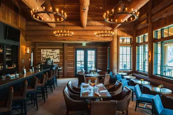 The interior of a rustic-chic dining area, featuring wooden log walls, elegant circular chandeliers, a sophisticated bar with stools, and various dining tables with chairs and banquettes, all illuminated by large windows offering an exterior view.