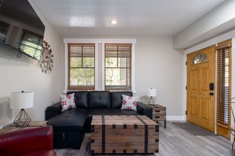 A comfortable lounge area within Town Pointe C106, featuring a black sectional sofa, a rustic wooden trunk coffee table, and natural light from large windows.