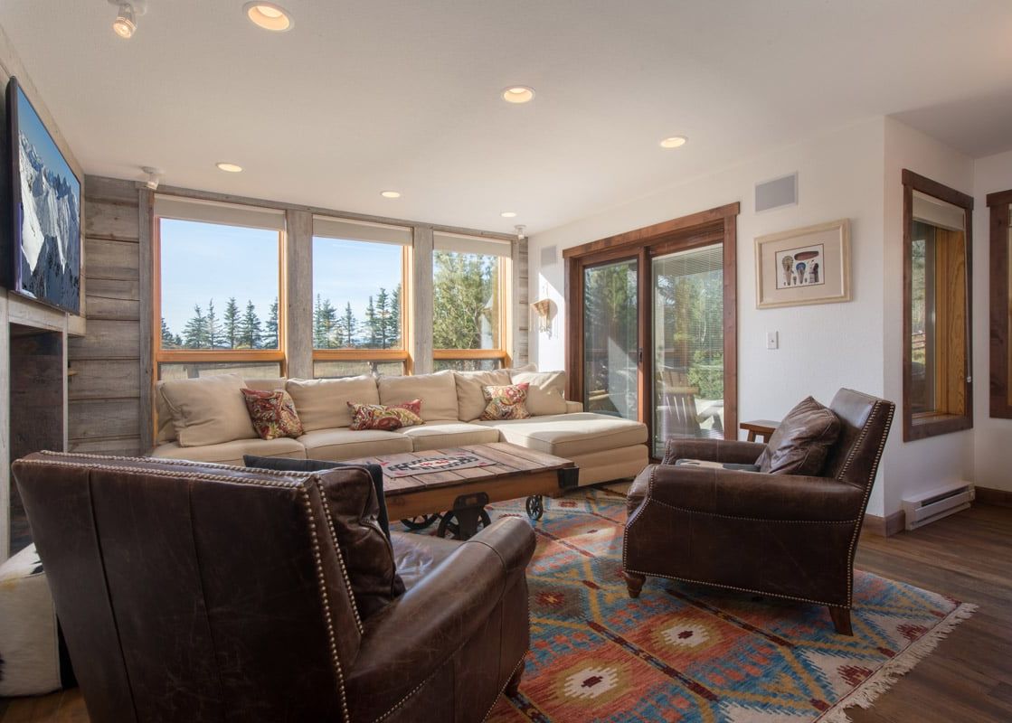 A spacious and inviting living room in Nez Perce D2 featuring a large L-shaped sectional sofa, two brown leather armchairs, a vibrant area rug, and large windows with a view of green trees and the sky.