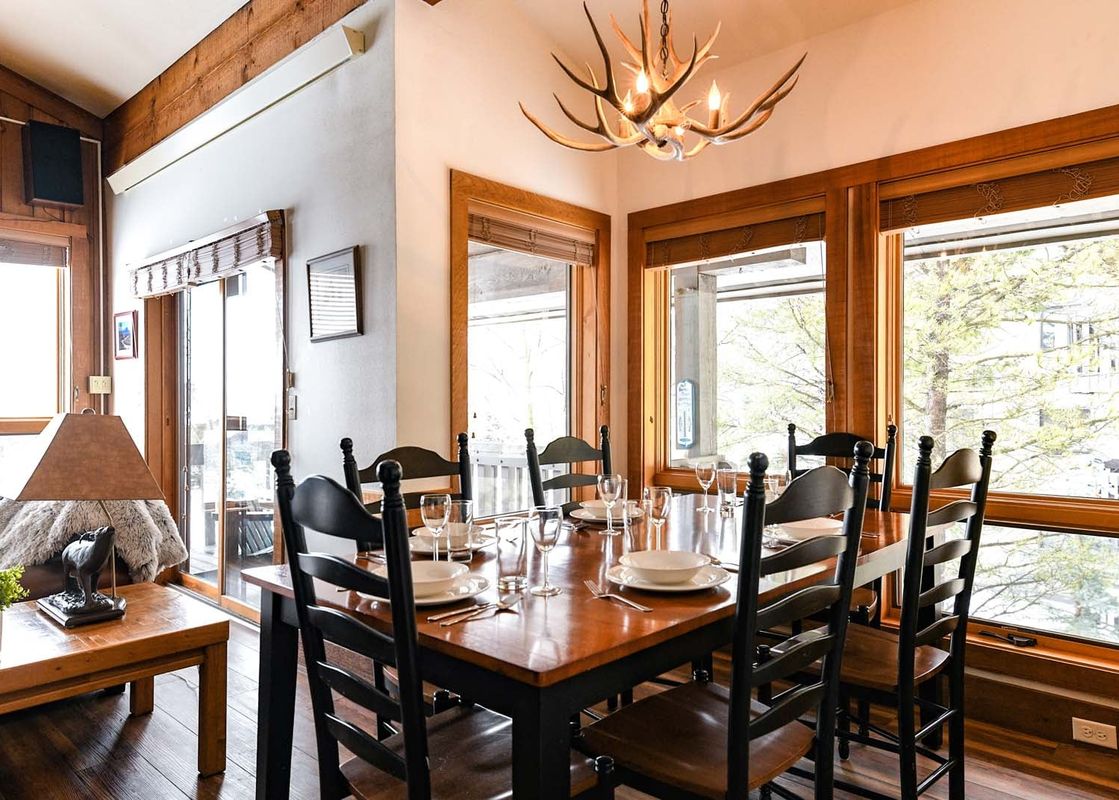 The wooden dining table and chairs in Nez Perce A6 under a rustic antler chandelier next to large bright windows.