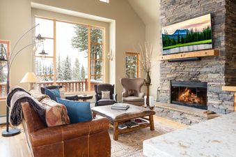 A cozy living room in Moonlight Mountain 38B Cowboy Heaven with a stone fireplace, comfortable leather seating, and large windows overlooking a snowy forest.