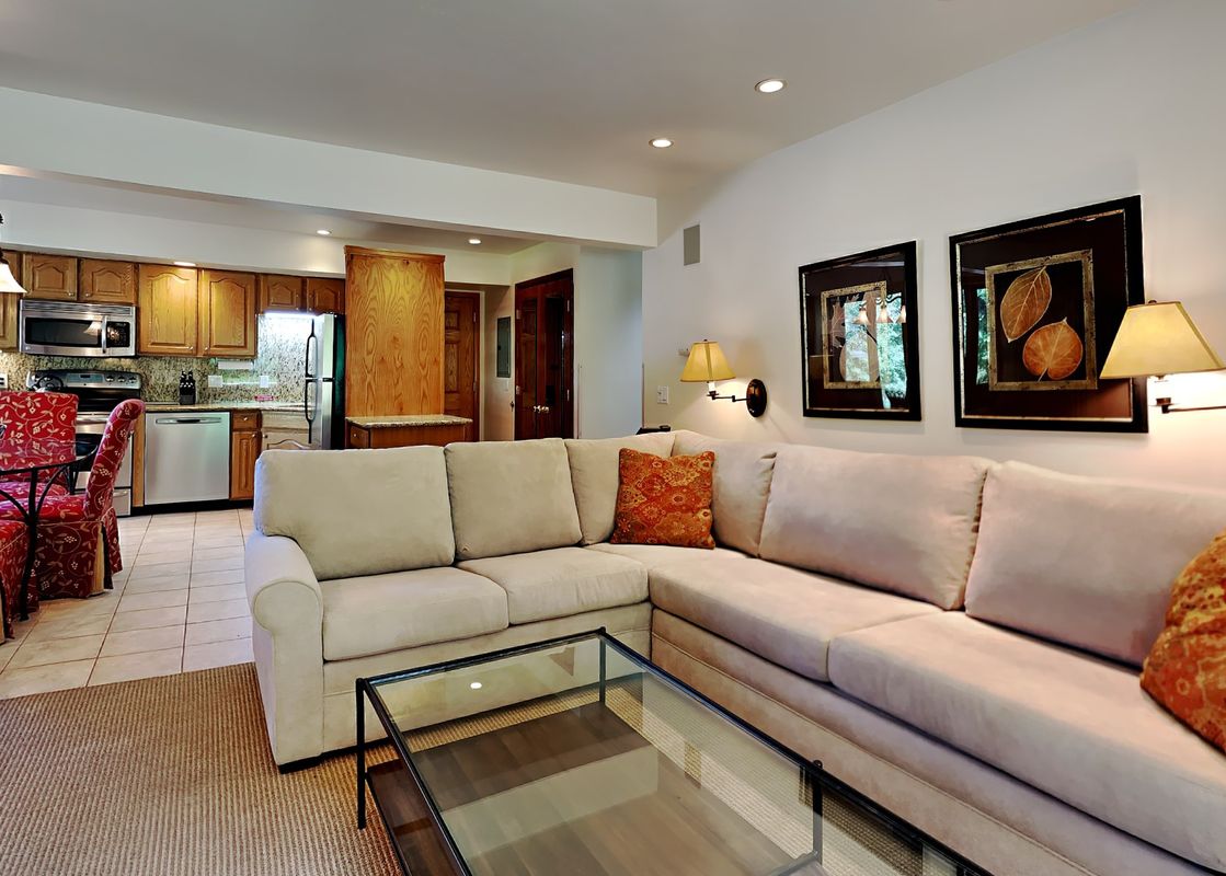 Open-plan living room with a large beige sectional sofa and a glass coffee table leading to the kitchen.